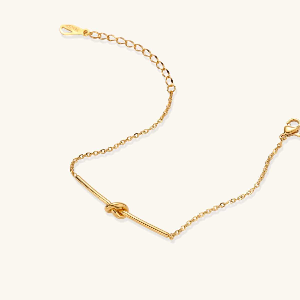 Delicate gold chain bracelet with minimalist bar and centered knot detail on cream background